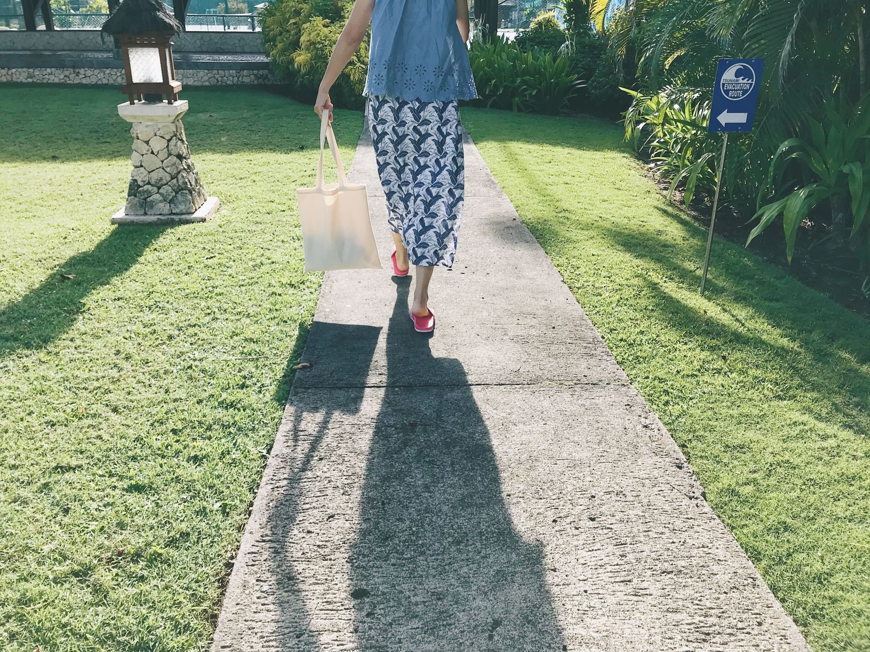 A woman with a tote bag walks down a sunlit garden path casting a long shadow.