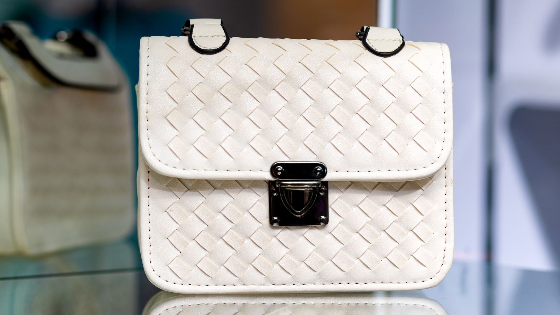 Stylish white leather handbag displayed on glass surface with intricate woven design and metal clasp.